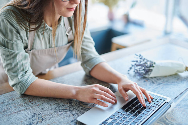 Mujer joven en delantal inclinándose sobre el teclado del ordenador portátil mientras navega por la red para obtener información
 - Foto, Imagen