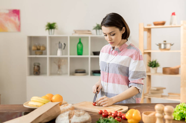 Young brunette woman in casualwear standing by kitchen table and cutting fresh tomatoes for salad - Foto, imagen