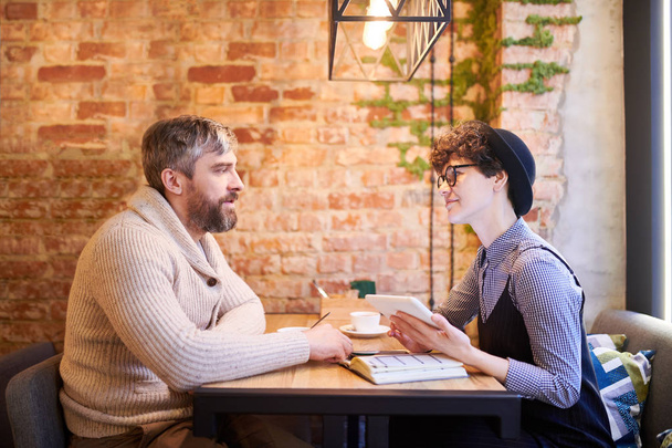 junges romantisches Paar in Freizeitkleidung, das im Café am Tisch sitzt und in aller Ruhe miteinander interagiert - Foto, Bild