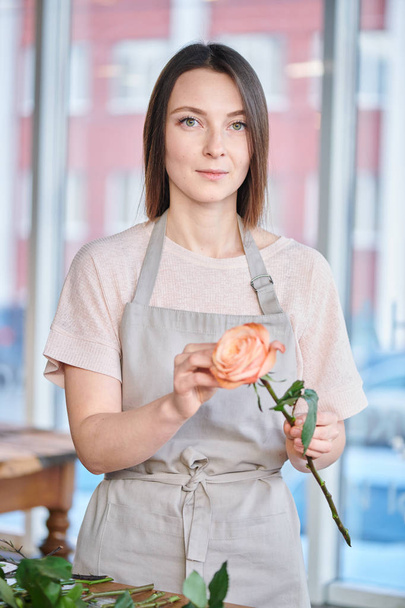 Όμορφη νεαρή γυναίκα στο λευκό μπλουζάκι και ποδιά κάνοντας τριαντάφυλλο τσαμπιά ενώ εργαζόταν στο ανθοπωλείο - Φωτογραφία, εικόνα
