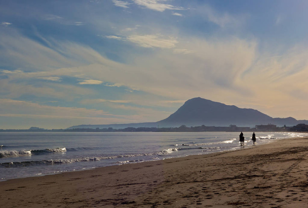 Waves roll on a sandy beach with horse riders. Clouds formation over mountains and the beach of Mediterranean Sea near Denia, Valencia, Spain. - Photo, Image