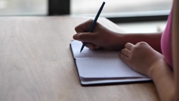 Una vista de cerca de una mujer afroamericana de raza mixta negra con una camisa rosa sentada en una mesa de comedor de madera y escribiendo una nota en su bloc de notas con un bolígrafo con las manos y el pecho en el marco
. - Imágenes, Vídeo