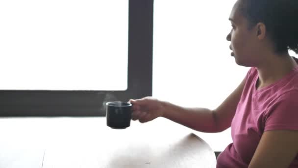 eine gemischte afrikanisch-amerikanische Frau im rosafarbenen T-Shirt holt sich eine heiße, dampfende Tasse Tee oder Kaffee von einem Esstisch mit walnussfarbenem Holztisch und Sonnenlicht, das aus einem Fenster drüber kommt. - Filmmaterial, Video