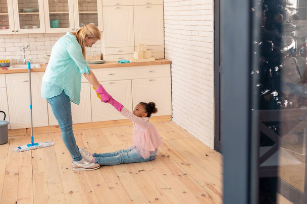 Cute funny girl sitting on the floor while mother cleaning the kitchen - 写真・画像