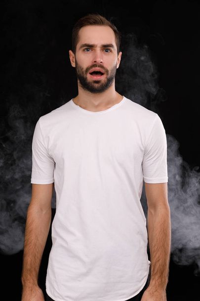Young guy in a white T-shirt on a black background with smoke - 写真・画像