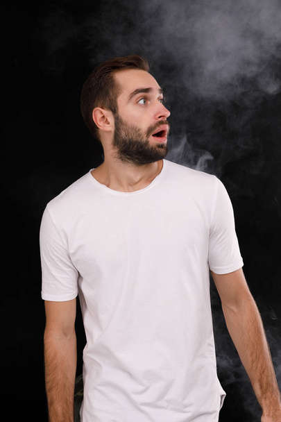 A young guy in a white T-shirt on a black background with smoke looks to the side - Photo, image