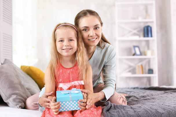 Schattig klein meisje met haar moeder en cadeau doos zittend op bed thuis - Foto, afbeelding