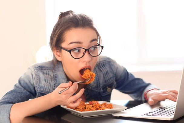 Young woman eating delicious pasta while using laptop indoors - Фото, зображення