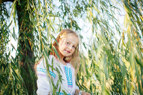 Cute blonde girl in white national dress with embroidery poses at the green tree at the spring festival - Φωτογραφία, εικόνα
