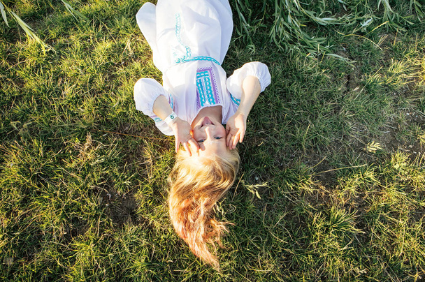 schattig meisje Blond in witte nationale jurk is liggend op het gras en lachen om het festival van de lente - Foto, afbeelding