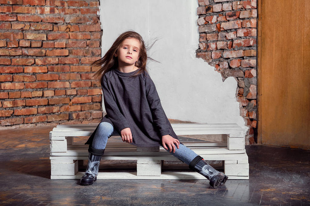 Little kid girl model posing fashionable in casual stylish clothes, gumboots. Fashion child sitting pose. studio background, brick wall.Shop youth, advertisement. - Photo, image