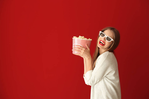Beautiful young woman with cup of popcorn and 3D cinema glasses on color background - Fotó, kép