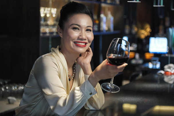 beautiful cheerful Asian businesswoman in light blouse relaxing with glass of wine in bar and looking at camera smiling on blurred background - Foto, Imagen
