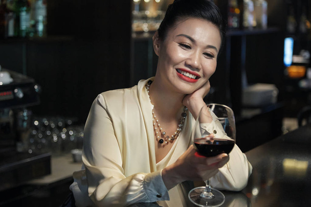 Zrelaksowany wesoły Asian kobieta elegancka bluzka i naszyjnik patrząc na kieliszek czerwonego wina w barze - Zdjęcie, obraz