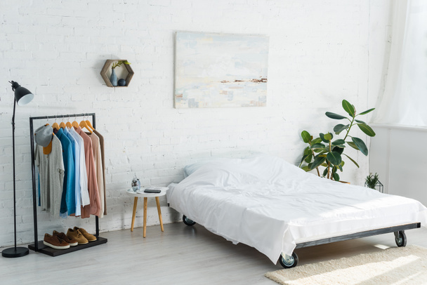 bed, clothes rack with shirts, plant, floor lamp and coffee table in bedroom - Photo, Image