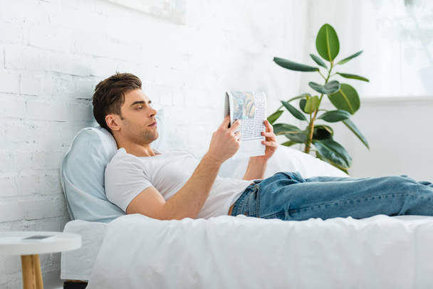 handsome man in white t-shirt and jeans lying on bed and reading newspaper in  bedroom - Foto, Imagem
