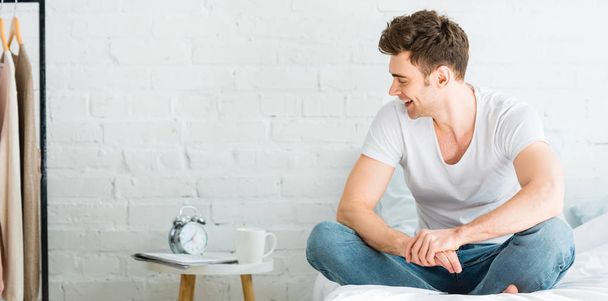 panoramic shot of handsome man in white t-shirt and jeans sitting on bed and smiling at home - Fotoğraf, Görsel