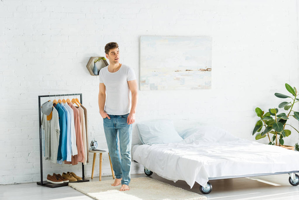 knappe man in wit t-shirt en jeans permanent in de buurt van bed en kleding rek in slaapkamer - Foto, afbeelding