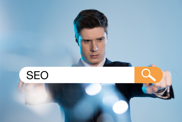 handsome businessman in suit pointing at search bar with seo letters in front on blue background - Foto, afbeelding