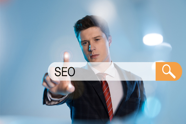 confident businessman in suit pointing with finger at search bar with seo letters in front on blue background - Photo, Image