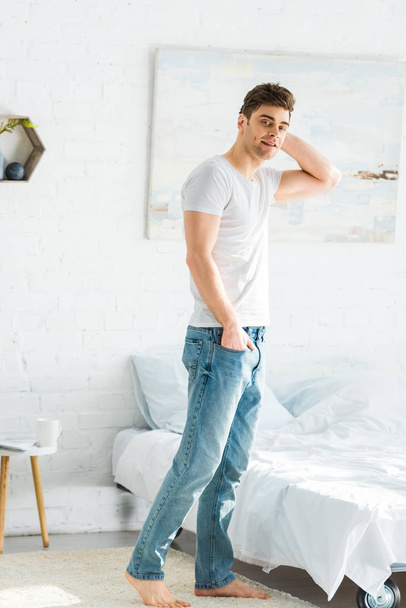 handsome man in white t-shirt and jeans standing near bed at home - Foto, Imagem