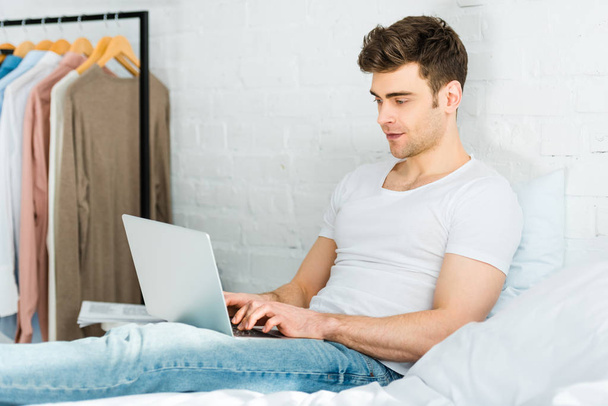 man in wit t-shirt en jeans zittend op bed en typen op laptop in slaapkamer - Foto, afbeelding