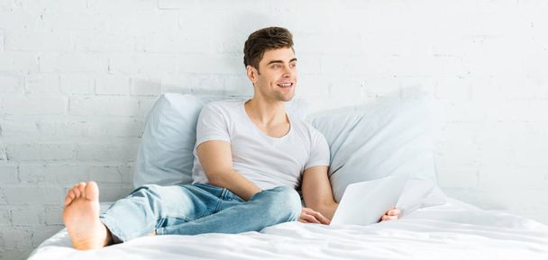 panoramic shot of handsome man in white t-shirt and jeans sitting on bed with laptop in bedroom - Photo, image