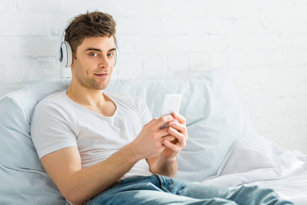 handsome man in white t-shirt and jeans sitting on bed with smartphone and listening music in bedroom - Photo, Image