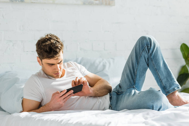 handsome man lying on bed and using black smartphone at home - Fotoğraf, Görsel