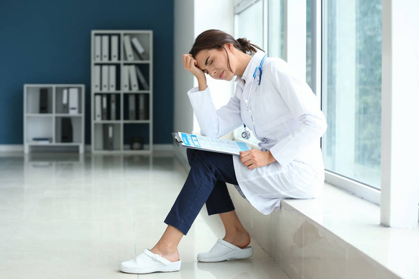 Doctora cansada sentada en el alféizar de la ventana en su consultorio
 - Foto, imagen