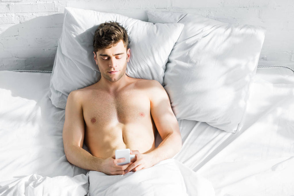 top view of handsome man with bare torso lying on bed with smartphone at home - Fotografie, Obrázek