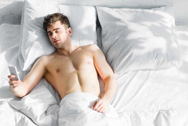 красивый мужчина с голым туловищем лежит на кровати и использует смартфон дома
 - Фото, изображение