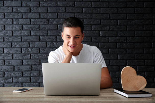 Mladý muž s online datování, zatímco sedí u stolu s notebookem proti cihlové zdi - Fotografie, Obrázek