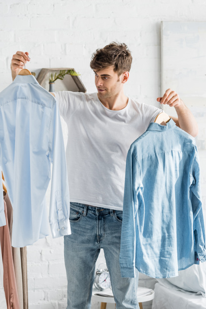 handsome man in white t-shirt holding shirts in bedroom - Foto, Imagen