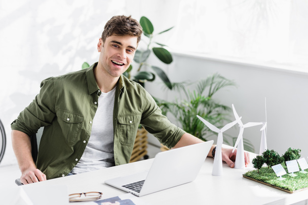 красивый архитектор сидит за столом с ноутбуком, ветряными мельницами, солнечными панелями модели на траве и улыбается в офисе
 - Фото, изображение