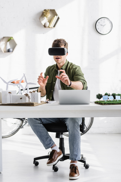 architect in zwarte VR headset aan tafel met laptop en windmolens, gebouwen, bomen, zonnepanelen modellen in kantoor - Foto, afbeelding