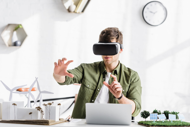 architect in virtual reality headset sitting at table with laptop and windmills, buildings, trees, solar panels models in office - Photo, Image