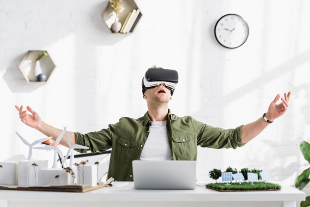 возбужденный архитектор в гарнитуре виртуальной реальности сидит за столом с ноутбуком и моделями в офисе
 - Фото, изображение