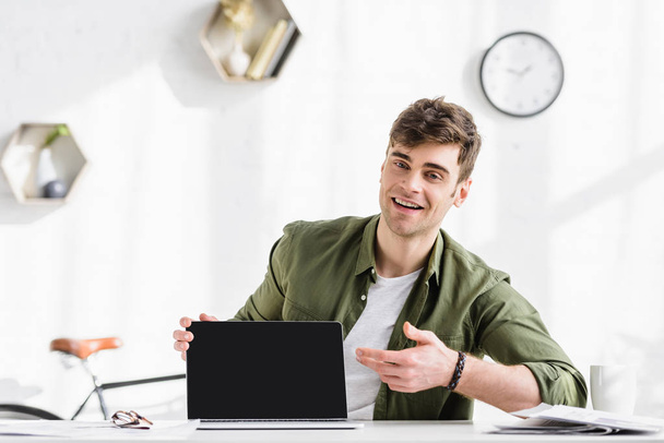 επιχειρηματίας σε πράσινο πουκάμισο, κάθεται στο τραπέζι και δείχνει με το χέρι σε φορητό υπολογιστή με κενή οθόνη στο γραφείο - Φωτογραφία, εικόνα