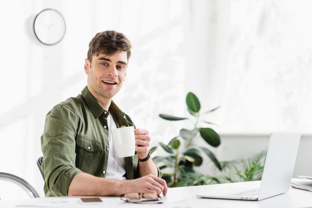 uomo d'affari in camicia verde seduto con tazza a tavola con computer portatile in ufficio
 - Foto, immagini