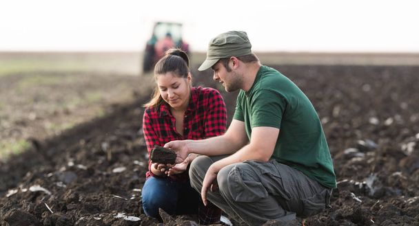 Jóvenes agricultores examinan la suciedad mientras tractor está arando campo
  - Foto, imagen