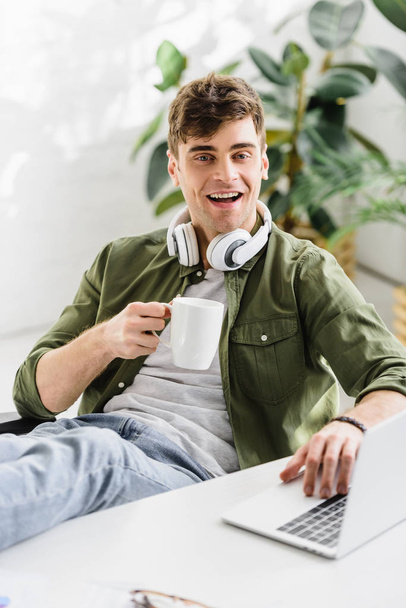 handsome businessman in headphones sitting, holding cup near table with laptop in office - Фото, зображення