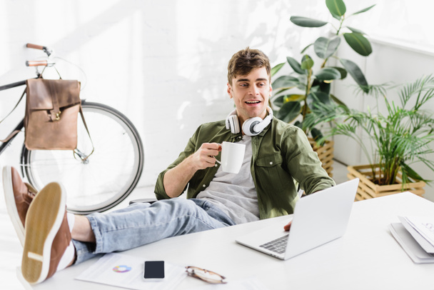 красивий бізнесмен в навушниках, сидячи, тримає чашку біля столу з ноутбуком в офісі
 - Фото, зображення