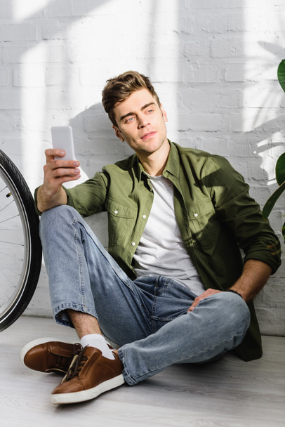 handsome man in green shirt holding smartphone, sitting near brick wall and bicycle in office - Zdjęcie, obraz