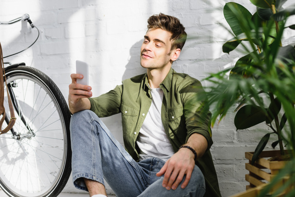 handsome man in green shirt holding smartphone, sitting near brick wall, bicycle and plants in office - Foto, Bild