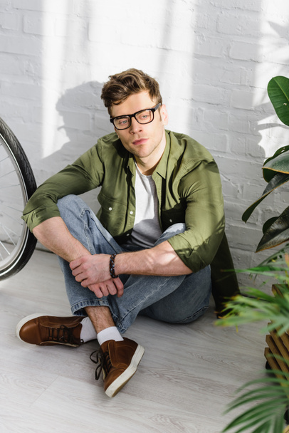 handsome man in green shirt, jeans and glasses sitting on floor near brick wall, bicycle and plants in office - Photo, Image