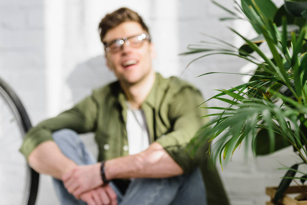 selective focus of man in green shirt, jeans sitting on floor near brick wall, and plants in office - Foto, Imagem
