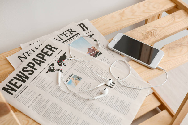 Teléfono móvil con auriculares y periódicos en estante de madera
 - Foto, Imagen