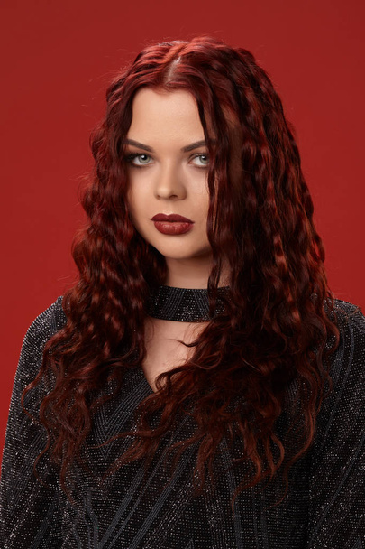Beautiful sexy woman is posing in a knitted dress on a red background. Evening makeup. Long, healthy, red curly hair. - Photo, Image