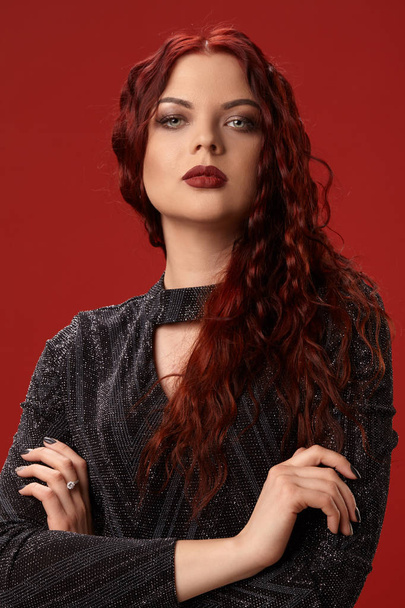 Belle femme sexy pose dans une robe tricotée sur un fond rouge. Maquillage de soirée. Cheveux longs, sains, bouclés rouges
. - Photo, image